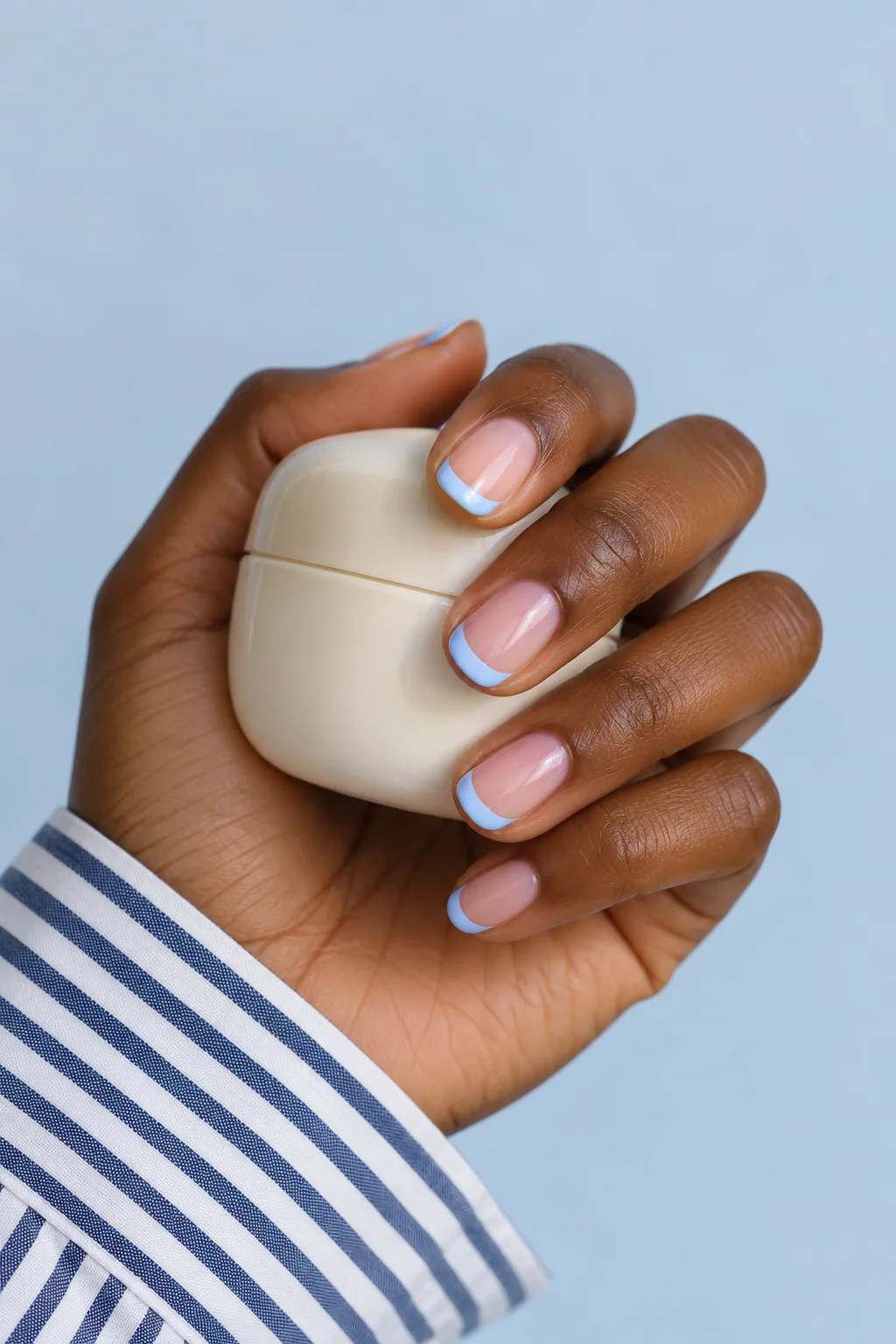 Powder-blue French tips on a hand holding an ivory jar above a striped cotton sleeve.